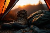 Image photoréaliste d'une paire de chaussures de randonnée robustes posées à côté d'un sac de couchage dans une tente, avec la lumière tamisée d'un coucher de soleil jetant un coup d’œil à travers la fermeture éclair, textures douces, éclairage atmosphérique chaleureux.